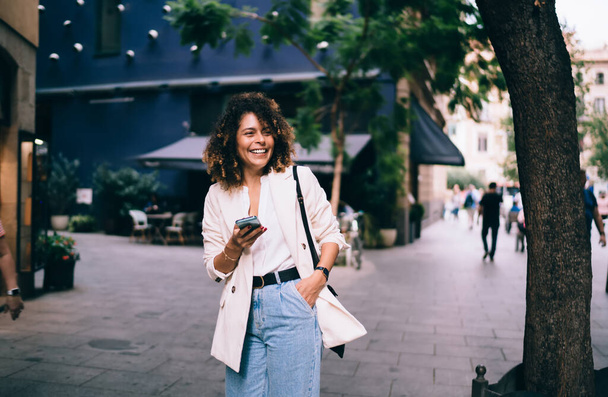 Joyful fashion blogger dressed in stylish streetwear laughing at touristic street rejoicing during solo vacations, funny hipster girl with modern cellular gadget smiling while walking in city - Foto, Bild