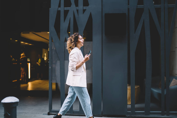 Side view of happy female tourist walking at street urban setting enjoying solo vacations for exploring world, trendy dressed millennial blogger with modern cellular device visiting modern city - Fotoğraf, Görsel