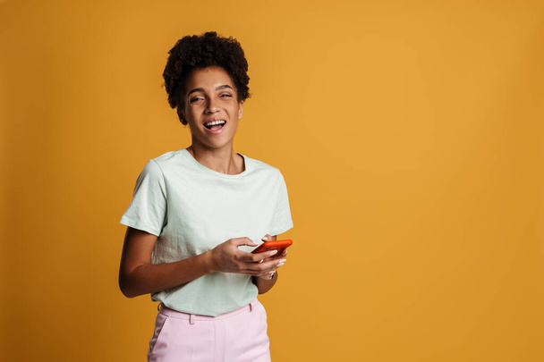Young black woman laughing while using mobile phone isolated over yellow background - Fotoğraf, Görsel