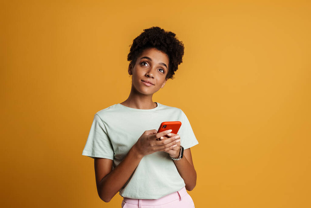Young black woman smiling while using mobile phone isolated over yellow background - Fotó, kép