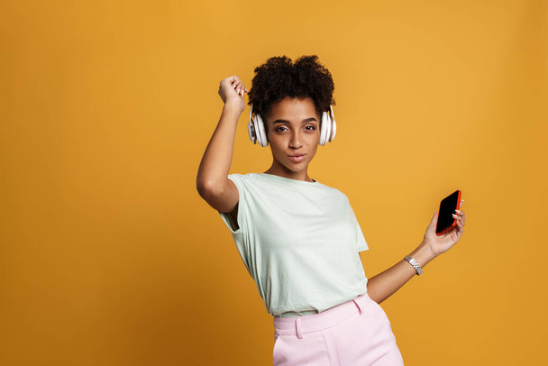 Young black woman listening music with headphones and dancing isolated over yellow background - Zdjęcie, obraz