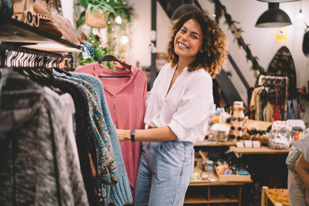 Portrait of joyful female customer enjoying weekend shopping in brand showroom, cheerful woman in trendy outfit buying stylish clothing and smiling at camera while posing near hangers with wear - Fotó, kép