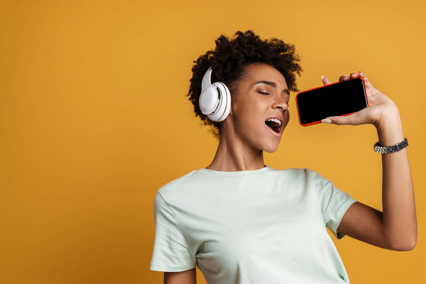 Молода чорна жінка слухає музику з навушниками та співає ізольовано на жовтому фоні
 - Фото, зображення