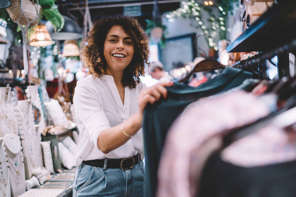Portrait of joyful female customer enjoying weekend shopping in brand showroom, cheerful woman in trendy outfit buying stylish clothing and smiling at camera while posing near hangers with wear - Φωτογραφία, εικόνα