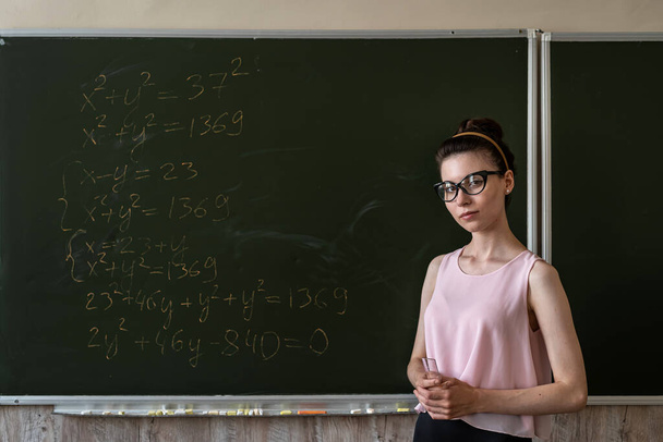 Młoda poważna nauczycielka wyjaśnia formuły matematyczne na uniwersytecie obok tablicy   - Zdjęcie, obraz