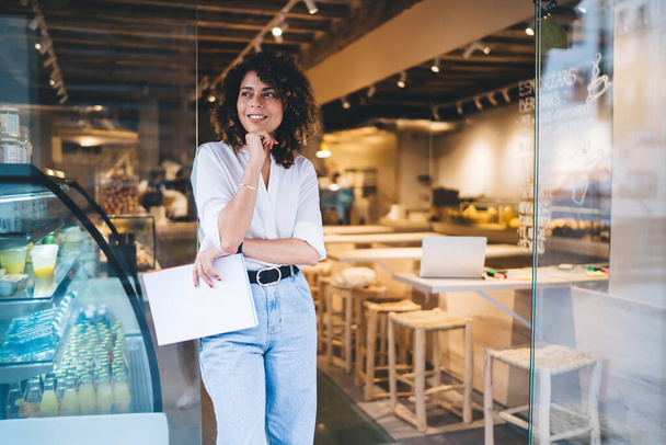 Happy female manager with menu in hand standing in doorway of local cafeteria and smiling, pretty Caucaisan small owner enjoying business lifestyle for working with franchise takeaway industry - 写真・画像