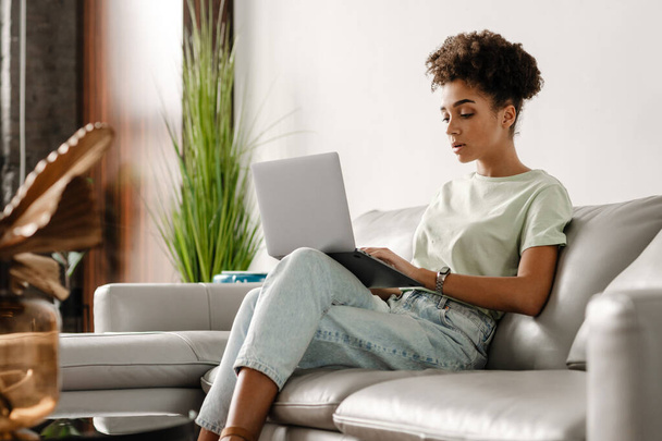 Young black woman working with laptop while sitting on couch at home - Zdjęcie, obraz