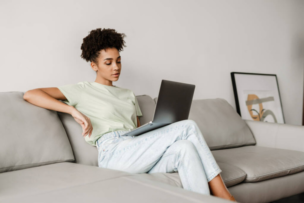 Young black woman working with laptop while sitting on couch at home - Foto, Imagen