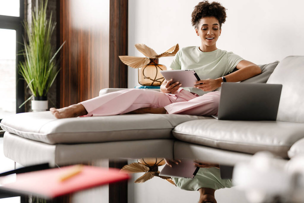 Young black woman using tablet computer while working with laptop at home - Foto, Bild