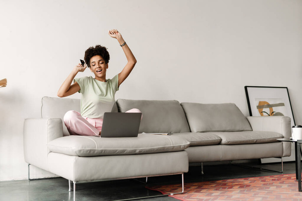 Young black woman gesturing and using credit card while working with laptop at home - Photo, image