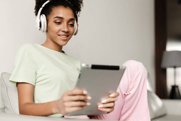Young black woman in headphones using tablet computer while sitting on couch at home - Foto, Imagem