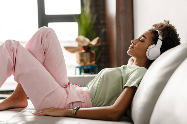 Young black woman listening music with headphones while sitting on couch at home - Φωτογραφία, εικόνα