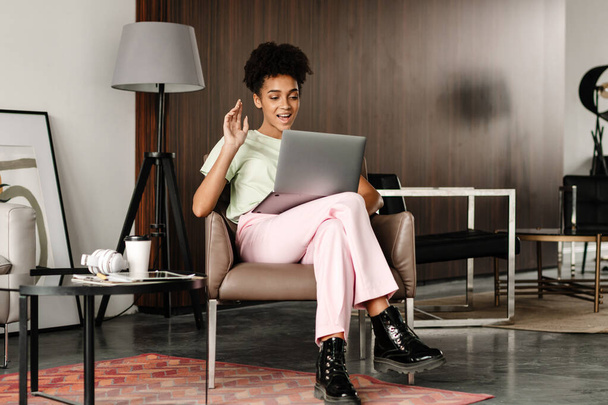 Young black woman gesturing and using laptop while sitting on armchair at home - Фото, зображення