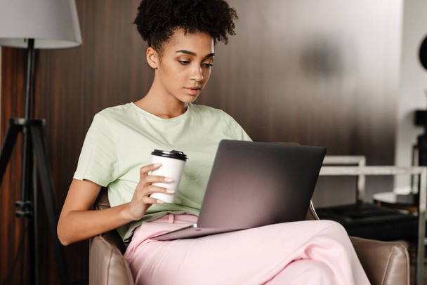 Νεαρή μαύρη γυναίκα πίνει καφέ ενώ εργάζεται με φορητό υπολογιστή στο σπίτι - Φωτογραφία, εικόνα