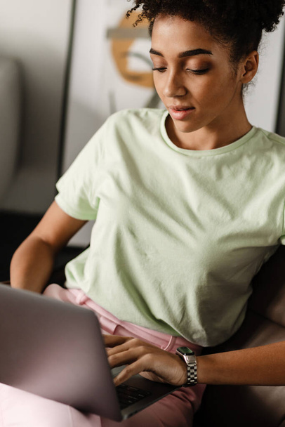 Young black woman working with laptop while sitting on armchair at home - Photo, image