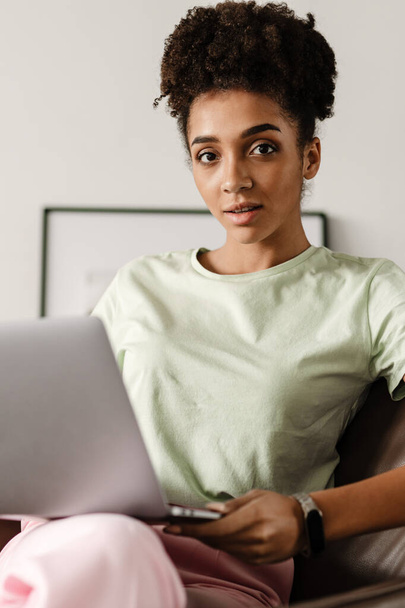 Mujer negra joven que trabaja con el ordenador portátil mientras está sentado en el sillón en casa - Foto, Imagen