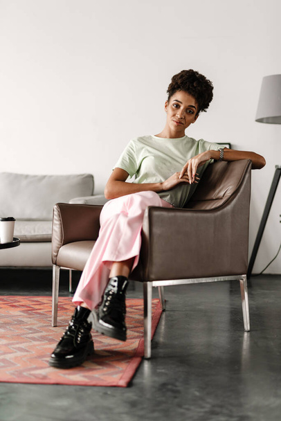 Young black woman smiling at camera while sitting on armchair at home - Фото, изображение