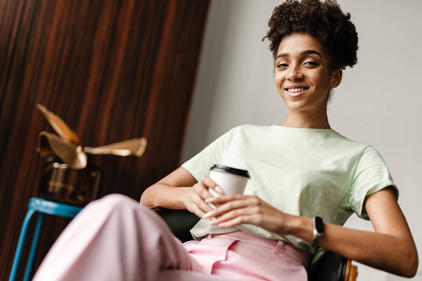 Young black woman smiling and drinking coffee while sitting on armchair at home - Foto, imagen
