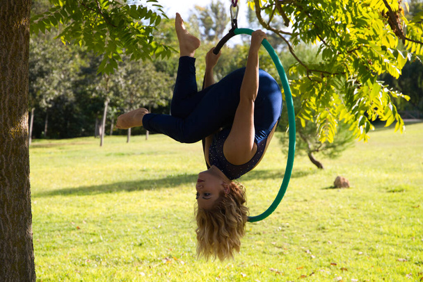 Mujer rubia y joven atleta acrobat gimnasta realizando ejercicio aéreo en el aire libre anillo en el parque. Lithe mujer en traje azul realiza poses de artistas de circo bailando con caderas. - Foto, imagen