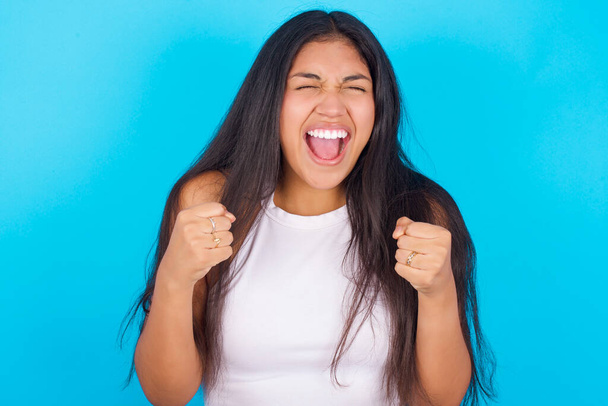 Молодая латиноамериканка в белой майке на синем фоне радуется успеху и победе, сжимая кулаки от радости, радуясь достижению цели и целей. Позитивные эмоции, чувства. - Фото, изображение