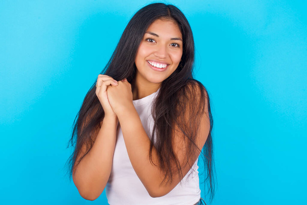 Dreamy Young hispanic girl wearing white tank top over blue background with pleasant expression, closes eyes, keeps hands crossed near face, thinks about something pleasant - Фото, изображение