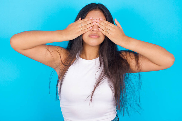 Young hispanic girl wearing white tank top over blue background  covering eyes with both hands, doesn't want to see anything or feeling ashamed. Human feelings reactions. - Photo, Image