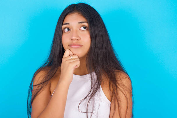 Portrait of thoughtful Young hispanic girl wearing white tank top over blue background  keeps hand under chin, looks away trying to remember something or listens something with interest. Youth concept. - Фото, изображение