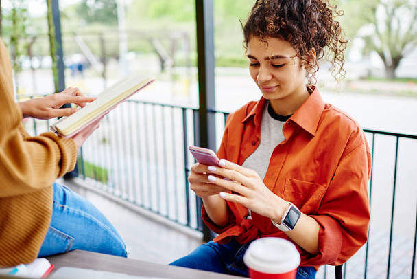 Crop ethnic female in casual clothes with curly hair sitting in cafe on street and using mobile phone for checking social network - Фото, изображение
