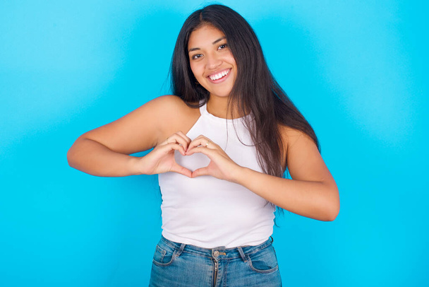 Mladá hispánská dívka nosí bílou tílko přes modré pozadí s úsměvem v lásce ukazující symbol srdce a tvar s rukama. Romantický koncept. - Fotografie, Obrázek