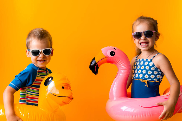 Two happy little children swimmer sport girl boy wear sun glasses swimsuit, Kid Inflatable swimming ring point to side away, isolated orange background. childhood, hot summer vacation hobby concept - Fotoğraf, Görsel