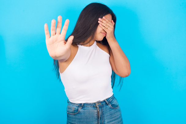 Jong Latijns-Amerikaans meisje draagt witte tank top over blauwe achtergrond bedekt ogen met palm en doen stop gebaar, probeert te verbergen. Kijk niet naar mij, ik wil niet zien, me schamen of bang zijn.. - Foto, afbeelding