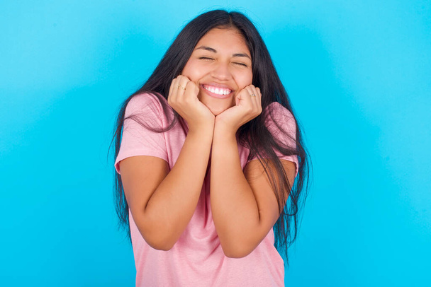 青い背景にピンクのTシャツを着た若いヒスパニック系の少女の肖像画が圧倒されます,顔の近くに閉じた目と手で興奮と幸せを表現. - 写真・画像