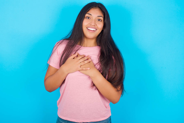 Šťastný úsměv Mladá hispánka v růžovém tričku přes modré pozadí má ruce na hrudi blízko srdce. Lidské emoce, skutečné pocity a pojetí výrazu obličeje. - Fotografie, Obrázek