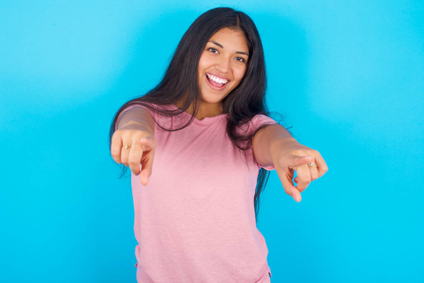 近くに驚きの若いヒスパニック系の少女ピンクのTシャツを着ての肖像画青の背景に2本の指でカメラを指していると言っ:私はあなたを選択! 、口を開けて検索. - 写真・画像
