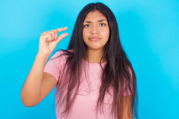 Upset Young hispanic girl wearing pink T-shirt over blue background shapes little gesture with hand demonstrates something very tiny small size. Not very much - Foto, Imagem