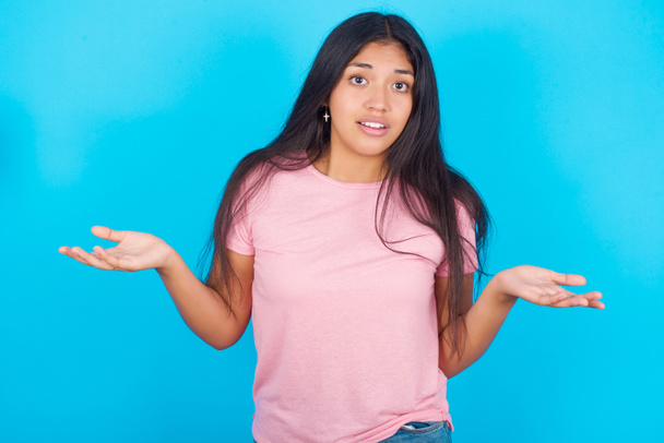 それで？傲慢な若いヒスパニック系の少女の肖像画ピンクのTシャツを身に着けている青い背景に手を横に笑みを浮かべて無関心な息をのむ、明らかに何かを伝える. - 写真・画像