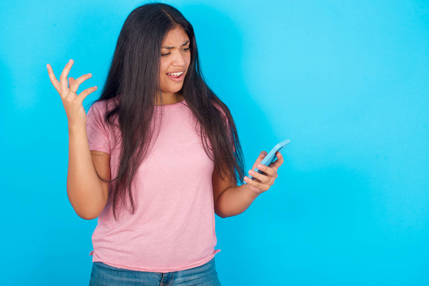 Photo of outraged annoyed Young hispanic girl wearing pink T-shirt over blue background holds cell phone, makes call, argues with colleague,  expresses negative emotions. People and anger. - Foto, Imagem