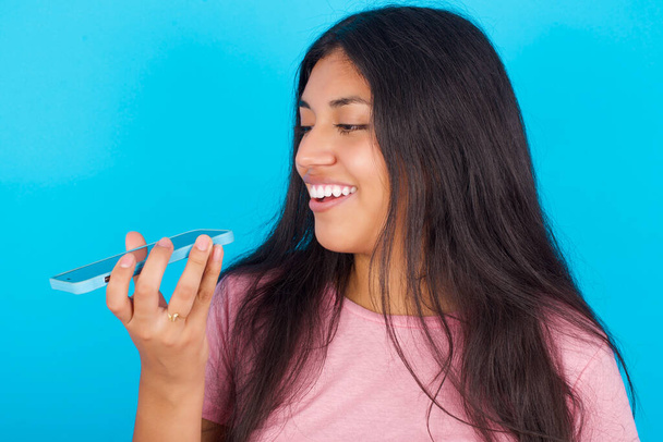 Usmívající se mladá hispánka v růžovém tričku nad modrým pozadím, posílající na svém chytrém telefonu hlasovou zprávu. Koncept komunikace a nových technologií. - Fotografie, Obrázek