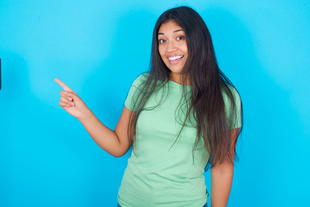 青い背景のポイントの上に緑のTシャツを着ている若いヒスパニック系の女の子は、コピー空白のスペースに脇。人々のプロモーションと広告のコンセプト - 写真・画像