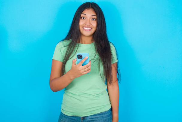 Fiatal spanyol lány zöld pólót visel kék háttér tartja a mobiltelefon a kezében, és örül a pozitív hír, használja a modern mobiltelefon - Fotó, kép
