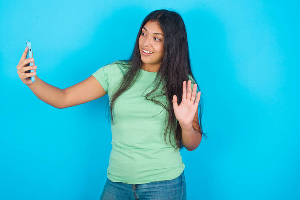 青い背景に緑色のTシャツを着た若いヒスパニック系の女の子は、現代の携帯電話を保持し、ハロージェスチャーでビデオ通話波の手のひらを作る。現代のテクノロジーの概念 - 写真・画像