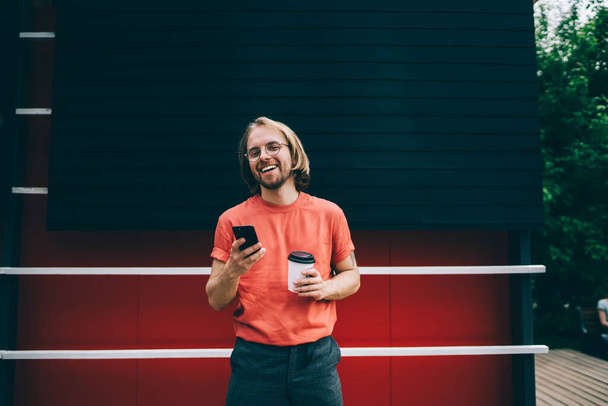 Young bearded modern hipster with long hair in red t shirt browsing mobile phone holding cup of takeaway coffee in city street - Фото, зображення