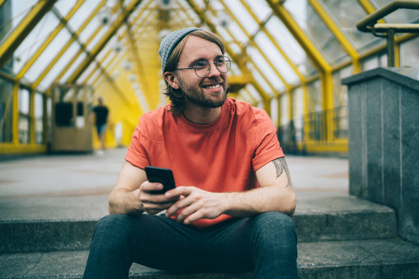 Молодий щасливий чоловік в капелюсі і окулярах посміхається і дивиться в сторону з мобільним телефоном в руці, сидячи перед скляним пішохідним переходом
 - Фото, зображення