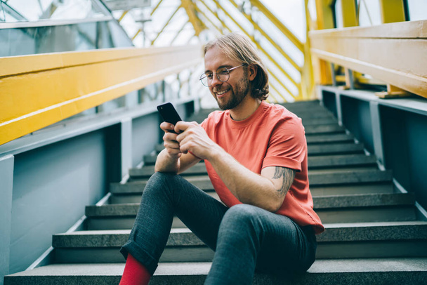 Carefree male in casual clothes and glasses using smartphone for browsing social media messaging with friend sitting on stairs indoors of urban building - 写真・画像