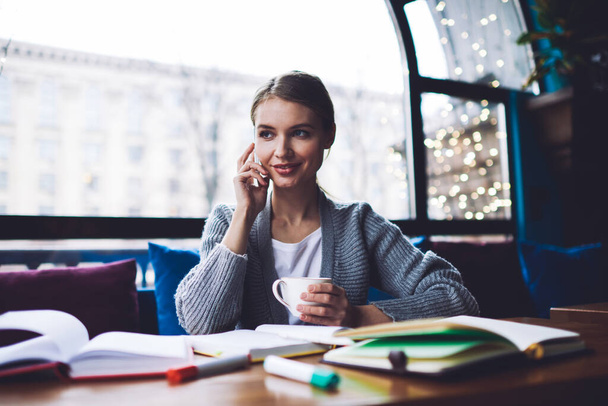 Jovem trabalhador remoto feminino feliz olhando para longe sentado à mesa com xícara de café e fazendo telefonema enquanto tem pausa no trabalho - Foto, Imagem