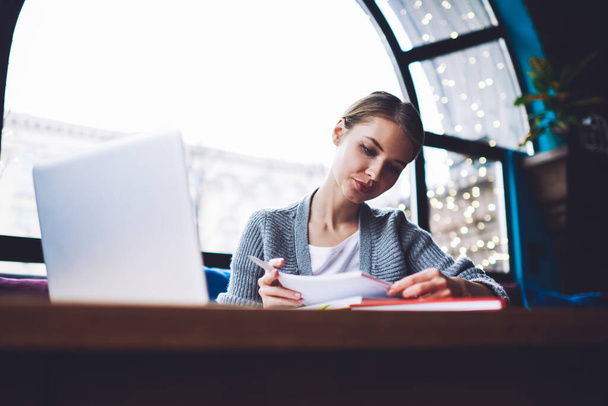 Freelancer feminino concentrado em roupas casuais lendo notas no caderno enquanto se senta à mesa com laptop e trabalha no café durante o dia - Foto, Imagem