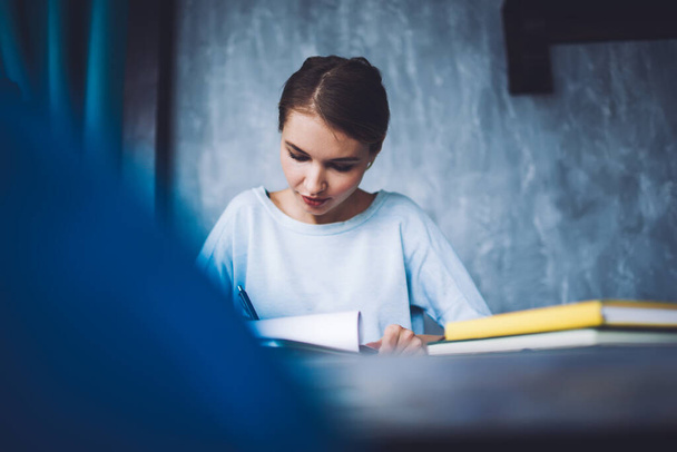 Estudiante femenina concentrada en ropa casual sentada a la mesa con bloc de notas y escribiendo información en hoja de papel mientras estudia - Foto, Imagen