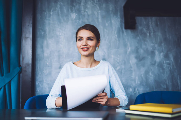 Cheerful female freelancer in casual outfit sitting at wooden table with notebooks while working in modern workspace and looking away - Foto, Imagem