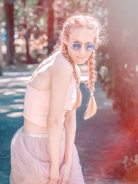 Smiling girl with two pigtails in a light pink summer outfit stands in a pose in a shady park - Foto, Imagem