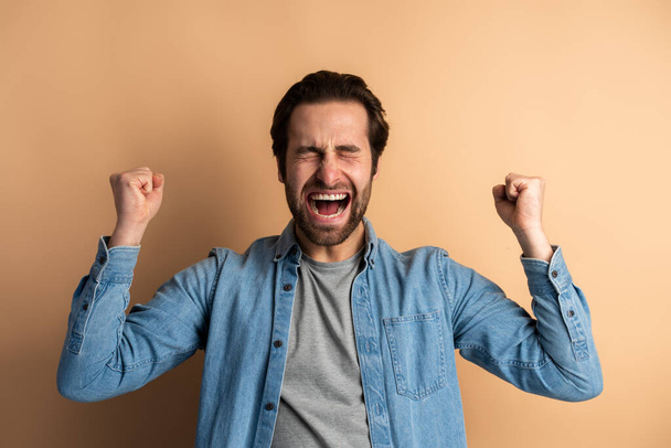 Portrait of ecstatic overjoyed handsome man in denim casual shirt expressing winning gesture with raised fists and screaming, celebrating victory. Indoor studio shot isolated on orange background  - Zdjęcie, obraz